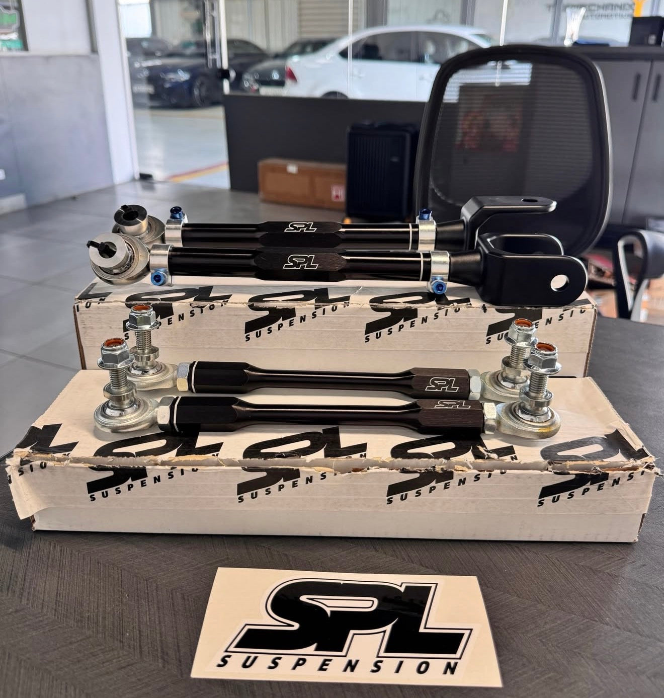 SPL Suspension front sway bar links and upper control arms displayed on their branded cardboard boxes on a dark tabletop, with an office chair and showroom cars in the background.